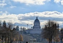 Washington Legislature Wants to Rein In Historic Landmarking to Spur Housing The domed Olympia Capitol Building stands in the distance with a bus stop with a passenger waiting in the foreground. A dogwalker also mosey on the lawn.