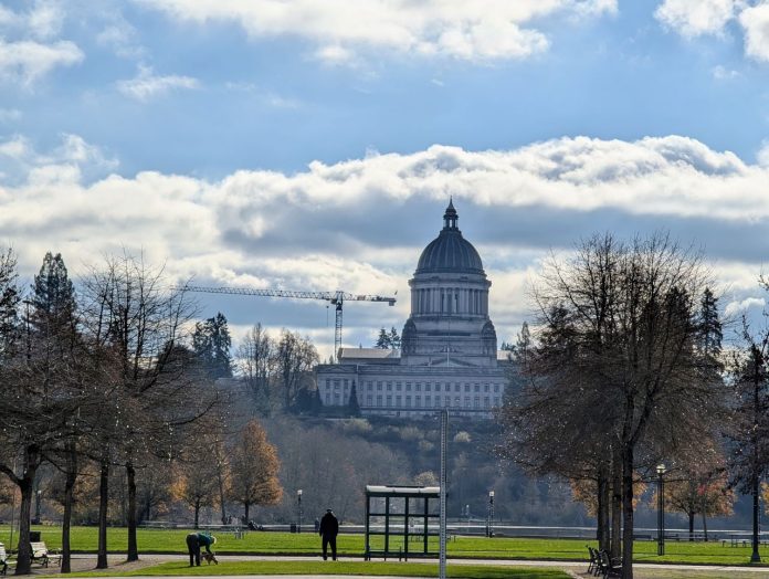Olympia Capitol dome by Doug Trumm 20241129 The domed Olympia Capitol Building stands in the distance with a bus stop with a passenger waiting in the foreground. A dogwalker also mosey on the lawn.