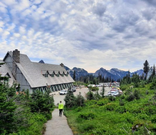 Op-Ed: Transit in Rainier National Park Would Ensure Paradise for Everyone A women walks down the trail to Paradise Inn and the crowded parking lot adjoining it. A mountain ridge and cirrus clouds are on the horizon.