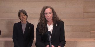 Davison is a white woman with curly hair in a dark blazer. She stands at a lectern in city hall with Sara Nelson behind her.