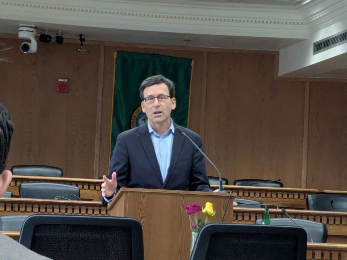 Bob Ferguson at Legislative Preview Day - Amy Sundberg_20250109 Ferguson stands at the lectern wearing a navy suit and glasses in a Capitol board room.