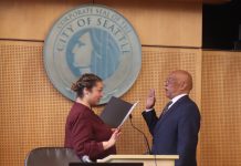 Seattle Council Appoints Mark Solomon to Fill District 2 Vacancy Solomon is a bald black man wearing eyeglasses and a dark suit. He stands in front of the Seattle City seal with his hand raised for his swearing in.