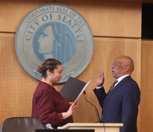 Seattle Council Appoints Mark Solomon to Fill District 2 Vacancy Solomon is a bald black man wearing eyeglasses and a dark suit. He stands in front of the Seattle City seal with his hand raised for his swearing in.