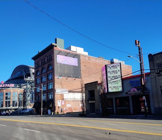 Seattle Council Sharply Divided Over Allowing Housing in Stadium District A photo from SoDo, with T-Mobile Park in the background. An older brick building stands next to smaller, more run down storefronts with a large wide road in the foreground