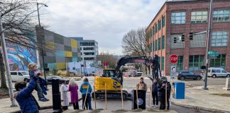 Bell Street is written into the pile of sand that was used in the ceremonial groundbreaking shoveling. A man hoists his child and officials mill about after the ceremony.