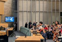 Public Meetings Privilege Housing Opponents — Here’s How to Fix It A women holds a baby with a toddler at her feet and speaks into the micro in Seatlte council chambers.