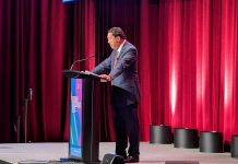 Harrell Calls Musk, Thiel ‘Smart Innovators,’ Tells Downtown Businesses He’s Fighting for Them Harrell stands at a lectern and looks down as his notes. He wears a grey suit and stands in front of a red curtain.