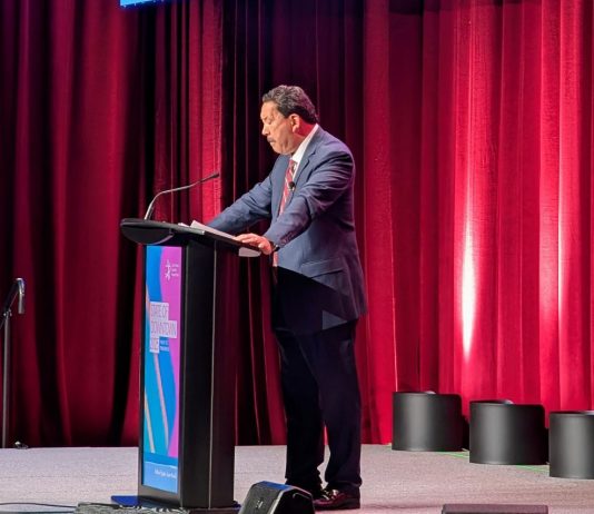 Harrell Calls Musk, Thiel ‘Smart Innovators,’ Tells Downtown Businesses He’s Fighting for Them Harrell stands at a lectern and looks down as his notes. He wears a grey suit and stands in front of a red curtain.