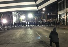 Op-Ed: Those Maimed by ‘Less Lethal’ Weapons Oppose Re-arming Police with Them A protester stands just to the right of a gaggle of protesters. A line of police officers approach.