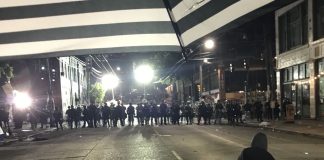 Op-Ed: Those Maimed by ‘Less Lethal’ Weapons Oppose Re-arming Police with Them A protester stands just to the right of a gaggle of protesters. A line of police officers approach.