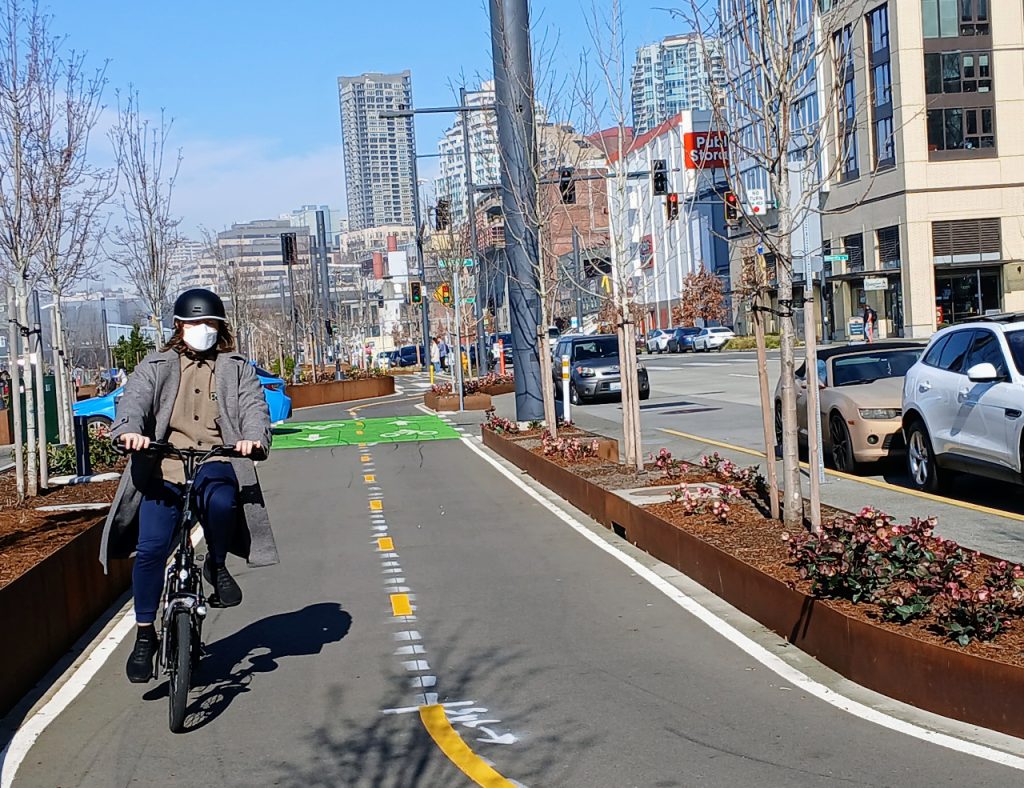 A person on a Brompton folding bike wearing a mask and biking south on Alaskan Way's bike lane near Seneca Street