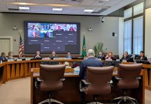 Sound Transit Hires Constantine as CEO in Unanimous Vote Constantine sits at fishbowl desk with a U-ring of seated boardmembers around him and a video screen with the members joining online.