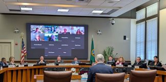 Constantine sits at fishbowl desk with a U-ring of seated boardmembers around him and a video screen with the members joining online.