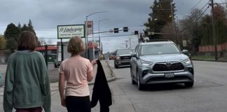 Two girls walk down 6th narrow sidewalk as cars zip by. One draps a jacket over her shoulder.