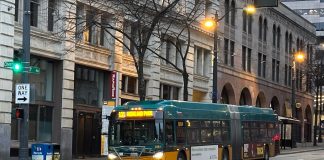 King County Council Wants Fewer ‘Ghost’ Buses and Better Data Collection A bus sports King County Metro's green and gold livery with masonry buildings in the background.