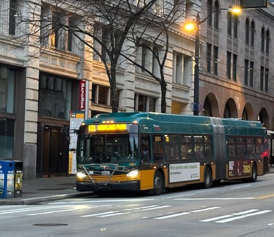 King County Council Wants Fewer ‘Ghost’ Buses and Better Data Collection A bus sports King County Metro's green and gold livery with masonry buildings in the background.