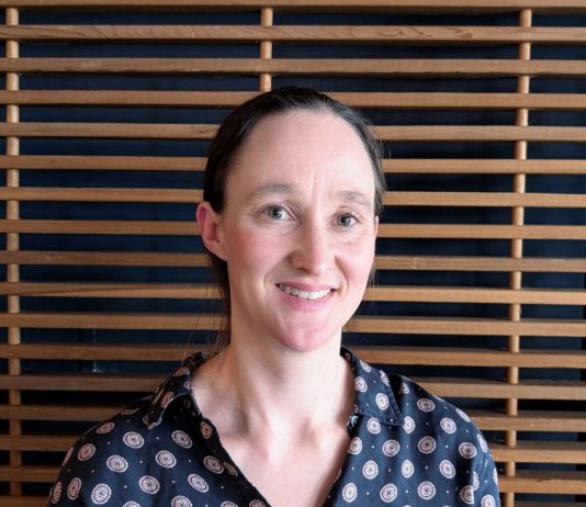Katie Wilson Jumps In Seattle Mayor Race as Progressive Coalition-Builder Katie wears a blue patterned blouse and stands in front of a wood slatted wall.