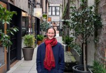 Wilson Pulls Within Two Points of Harrell in Friday Count Wilson wear a red scarf and a dark blue coast and stand in the Chophouse Row alley which is lined by potted plants and restaurant storefronts.