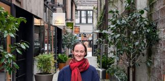Wilson wear a red scarf and a dark blue coast and stand in the Chophouse Row alley which is lined by potted plants and restaurant storefronts.