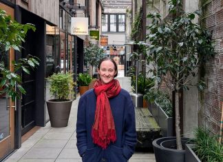 Wilson Pulls Within Two Points of Harrell in Friday Count Wilson wear a red scarf and a dark blue coast and stand in the Chophouse Row alley which is lined by potted plants and restaurant storefronts.