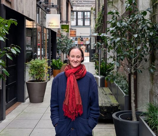 Wilson Pulls Within Two Points of Harrell in Friday Count Wilson wear a red scarf and a dark blue coast and stand in the Chophouse Row alley which is lined by potted plants and restaurant storefronts.