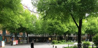 A quiet pedestrian street with a few people walking and plenty of tree canopy with a midrise residential building in the background.