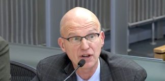 Torgelson wears glasses, a blue checkered tie and a dark suit. He's a bald White man.