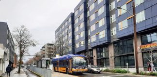 A travels down Roosevelt Way next to the protected bike lane. A large seven-story apartment building with a crawfish restaurant on the first floor is on the right.