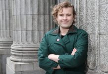 Ry Armstrong Lays Out Progressive Contrast Against Mayor Harrell Ry stands in front of a Greek column with their arms crossed