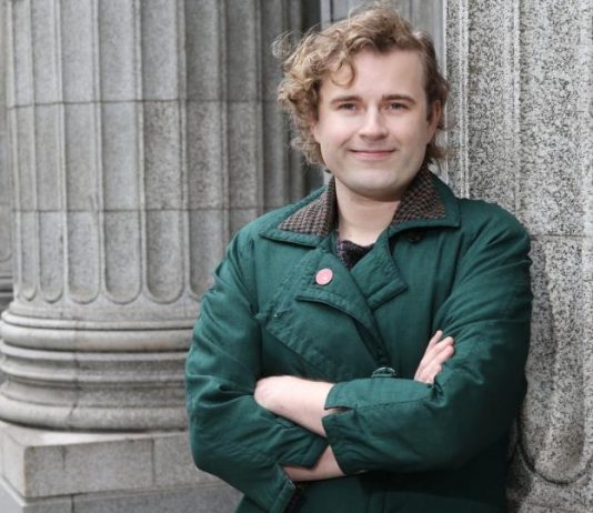 Ry Armstrong Lays Out Progressive Contrast Against Mayor Harrell Ry stands in front of a Greek column with their arms crossed