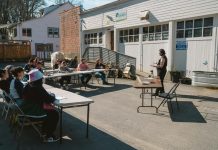 Op-Ed: Why Your City Needs a Tool Library A woman leads a lass which is sitting around folding tables in the parking lot of the tool library building, and its rows of garage doors.