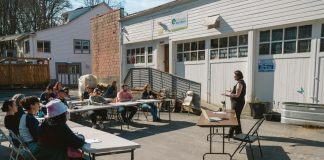 Op-Ed: Why Your City Needs a Tool Library A woman leads a lass which is sitting around folding tables in the parking lot of the tool library building, and its rows of garage doors.