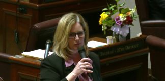 Cleveland hold the microphone on the floor and has blond hair and glasses. On the Annette Cleveland desk behind her is a bouquet of flowers.
