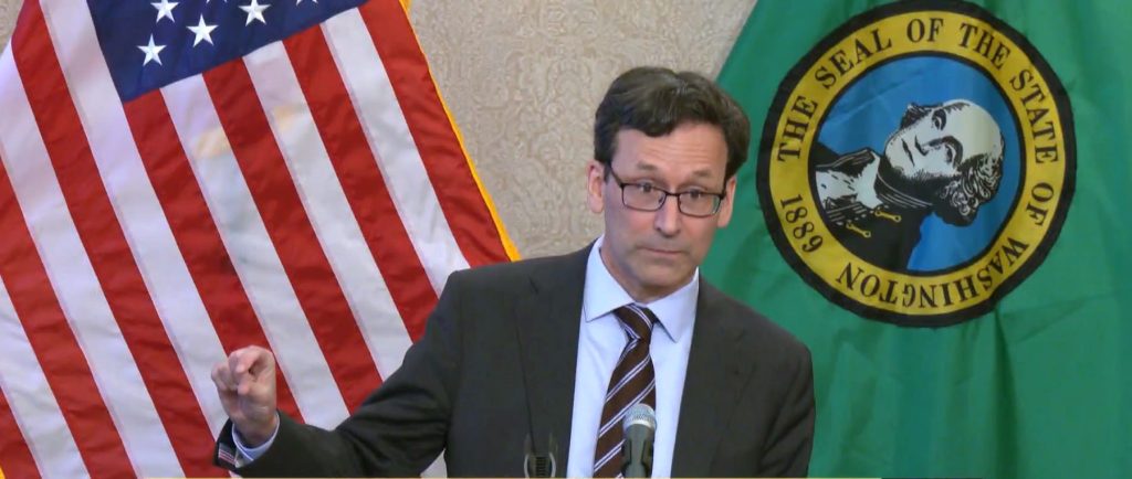 Ferguson wears a suit and glasses and gestures his figures with a little bit sign. He stands in front of the USA and Washington state flags.