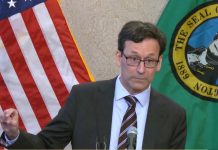 Ferguson Ignites Revenue Battle with State Democrats, Threatening Veto Ferguson wears a suit and glasses and gestures his figures with a little bit sign. He stands in front of the USA and Washington state flags.