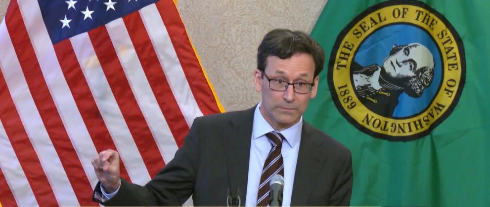 Ferguson wears a suit and glasses and gestures his figures with a little bit sign. He stands in front of the USA and Washington state flags.