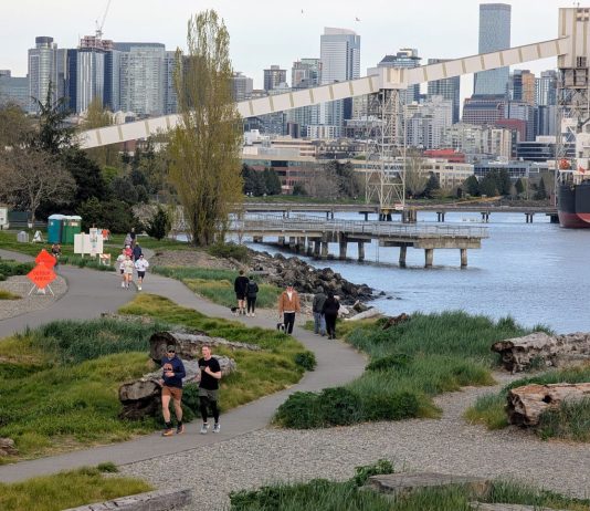 Elliott Bay Trail Upgrades Temporarily Close Waterfront Bike Path Orange signs warn of the bike path closure just east of Expedia Park. A dozen joggers and dog walkers crowd the waterfront path. The Seattle skyline is in the distance.