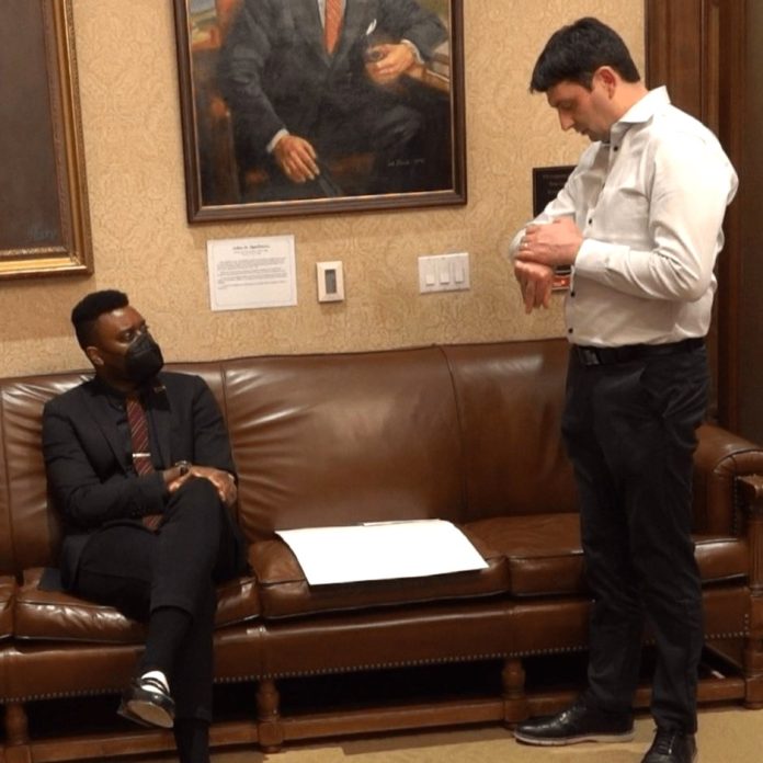 Shaun Scott and Ferguson Chief of Staff recrop Shane Esquibel answers a call coming through on his smartwatch, as Scott sits and looks in from the Governor's waiting room, adorned with official portraits of governors long past.