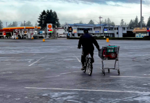 Bremerton Ponders Ending Parking Mandates to Spur Housing A person bikes pulling a shopping card alongside at a big empty parking lot outside a big box store.