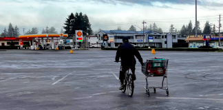 A person bikes pulling a shopping card alongside at a big empty parking lot outside a big box store.