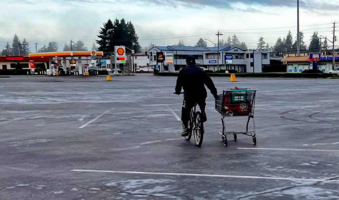 An empty parking lot A person bikes pulling a shopping card alongside at a big empty parking lot outside a big box store.