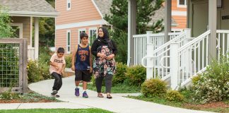 A mother and her two sons walk on a pedestrian path between a row of townhomes. She wears a headscarf and her older son wears a Lebron James jersey. The younger is on a skateboard.