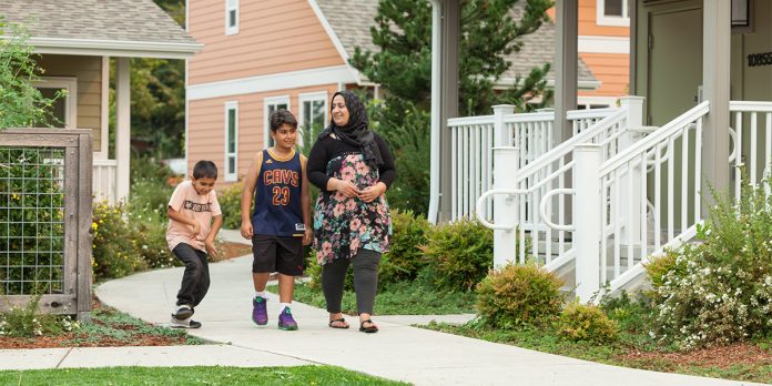 Family at King County Housing Authority site A mother and her two sons walk on a pedestrian path between a row of townhomes. She wears a headscarf and her older son wears a Lebron James jersey. The younger is on a skateboard.