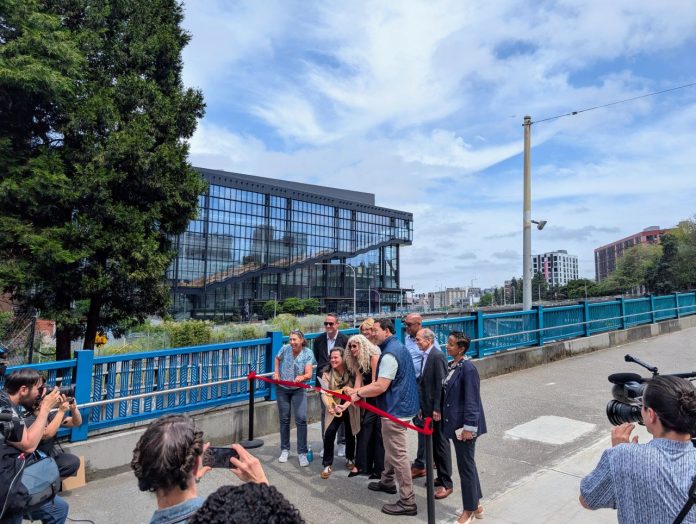 Harrell Pike Pine RibbonCutting by Trumm 20250611 Harrell holds ceremonial gold scissors to cut the red ribbon on a widen sidewalk next to a new protected bike lane on Pike Street on the I-5 overpass bridge to Capitol Hill. The new convention center is in the background.