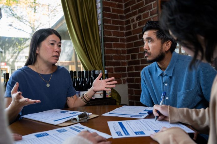 Jeanie Chunn at table via campaign An Asian woman with straight long black hair, Chunn sits at a table gesturing broadly with hands talking to the man seated next to her.