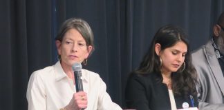 Seattle Council Candidates Spar at Mobility and Housing Forum Sara Nelson holds a microphone and wears a white button-up shirts. She sits a long table next to Alexis Rinck, who wears a black blazer and hoop earrings.