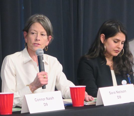 Seattle Council Candidates Spar at Mobility and Housing Forum Sara Nelson holds a microphone and wears a white button-up shirts. She sits a long table next to Alexis Rinck, who wears a black blazer and hoop earrings.