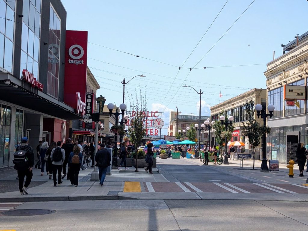 The iconic Pike Place Market sign is in the distance and umbrellas and patio furniture enlivens the middle of the pedestrianized street. The sidewalk is packed with people walking.