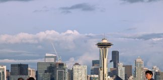 A group of young people stand by the railing at Kerry Point with the Space Needle and downtown skyscrapers looming in the distance.