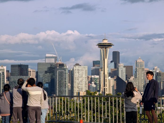 Seattle space needle skyline from Kerry Park_ Trumm 20250621 A group of young people stand by the railing at Kerry Point with the Space Needle and downtown skyscrapers looming in the distance.